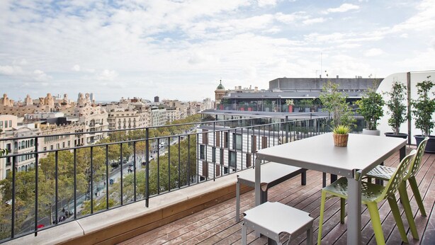 A balcony with a table and chairs, overlooking a cityscape with buildings and a clear sky. - Bonavista Apartments - Pedrera (Barcelona)