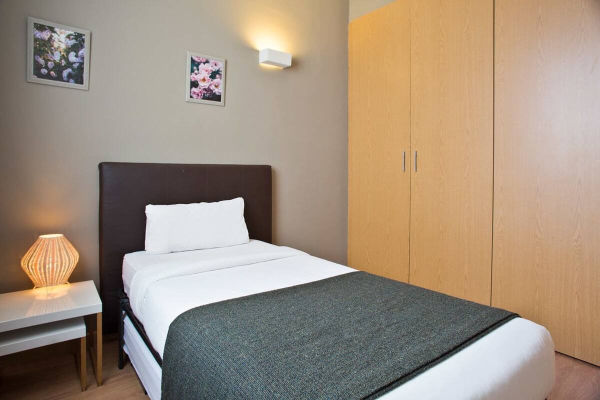 Un lit bien fait, avec une couette blanche et un plaid gris, une lampe de chevet et une armoire en bois à deux portes.