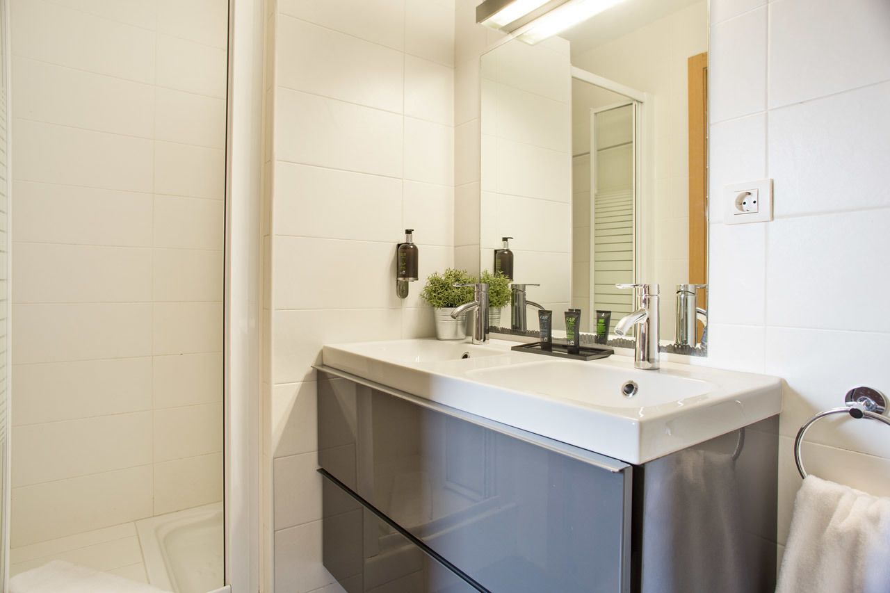 A modern bathroom with a white tiled wall, a large mirror, a white sink with a black faucet, and a small potted plant on the counter.