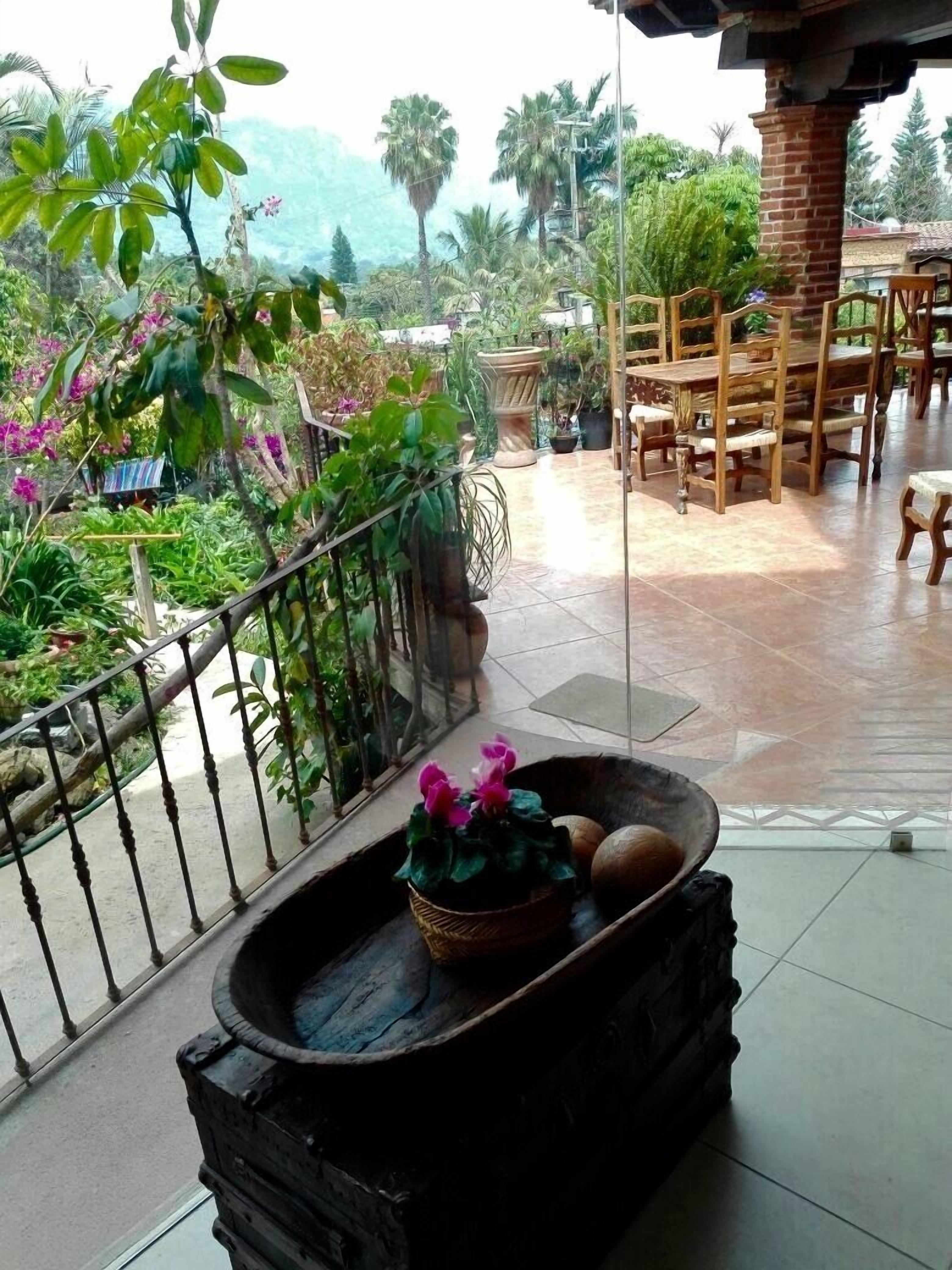 Un balcón con vistas a un jardín, una mesa y sillas de madera, y una maceta decorativa con flores y frutos.