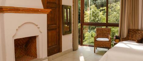 A bedroom with a fireplace, a large window with a view of greenery, a wicker chair, and a bed.