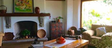 A living room with a fireplace, a wooden coffee table with books and a candle, a sofa, and a window with a view of greenery.