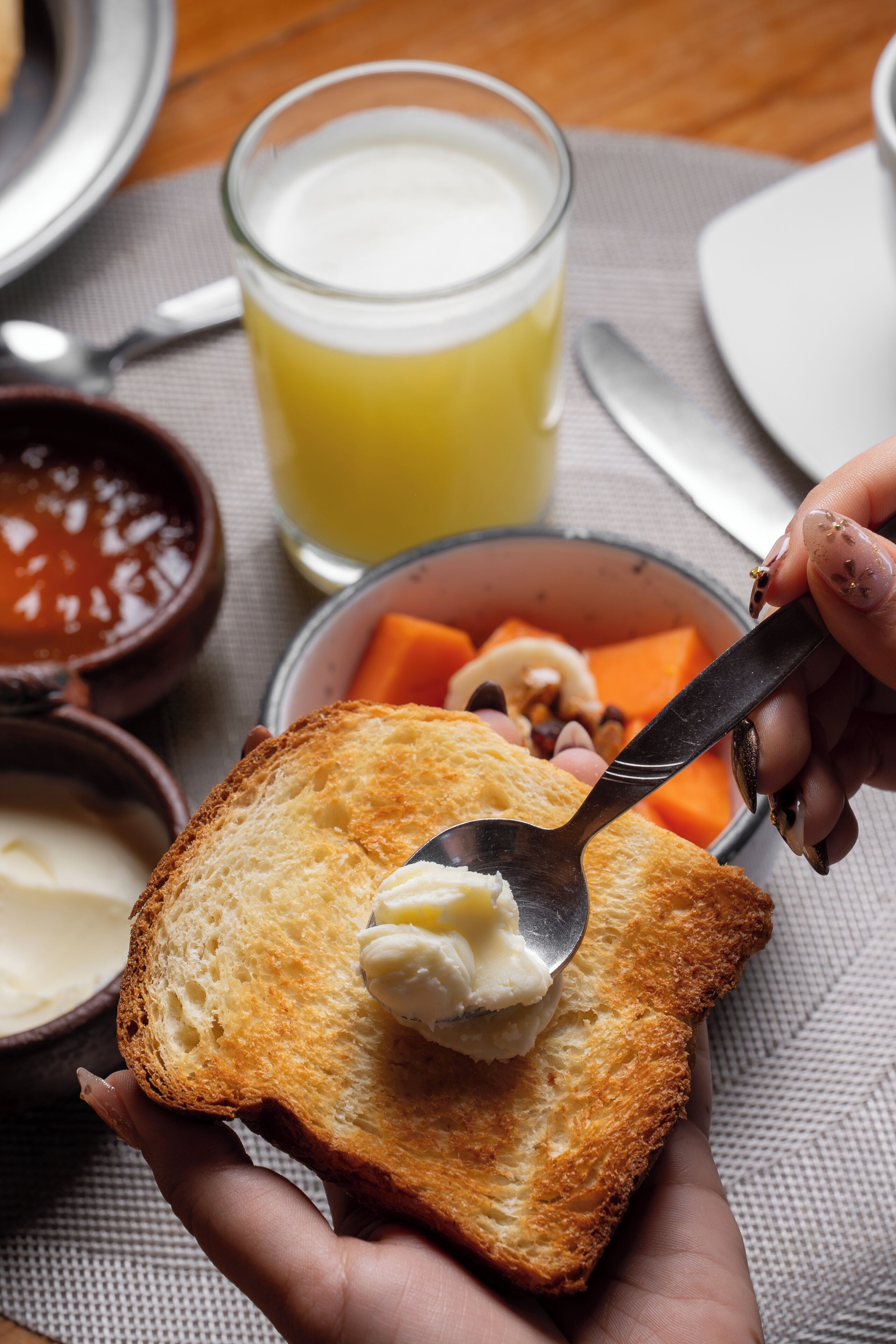 Una persona untando mantequilla en tostadas con una cuchara, junto a un vaso de jugo de naranja, cuencos de sopa y un plato de fruta.