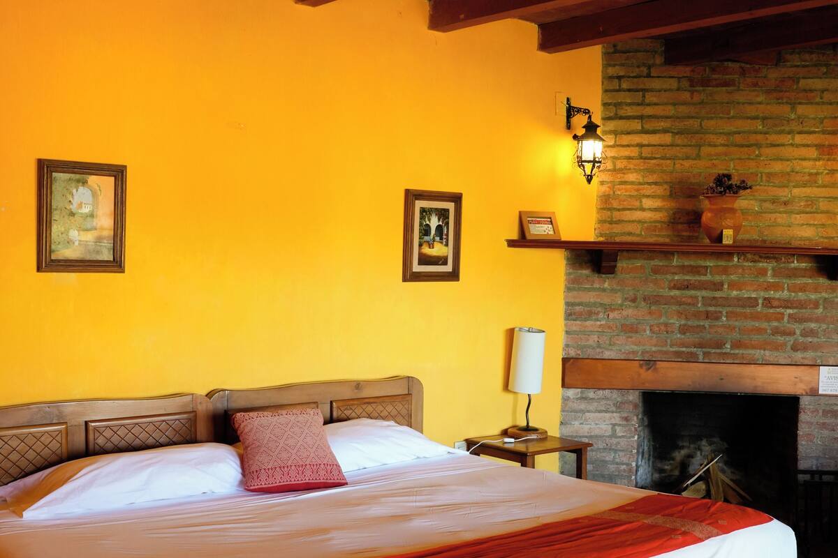 A room with a large bed, wooden headboard, a brick fireplace, and a yellow wall.