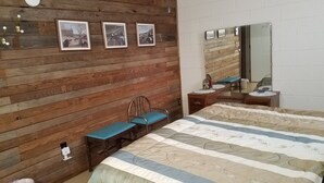 Una habitación con pared de madera, una cama con cubrecama a rayas, una silla, una mesita con espejo y tres cuadros enmarcados.