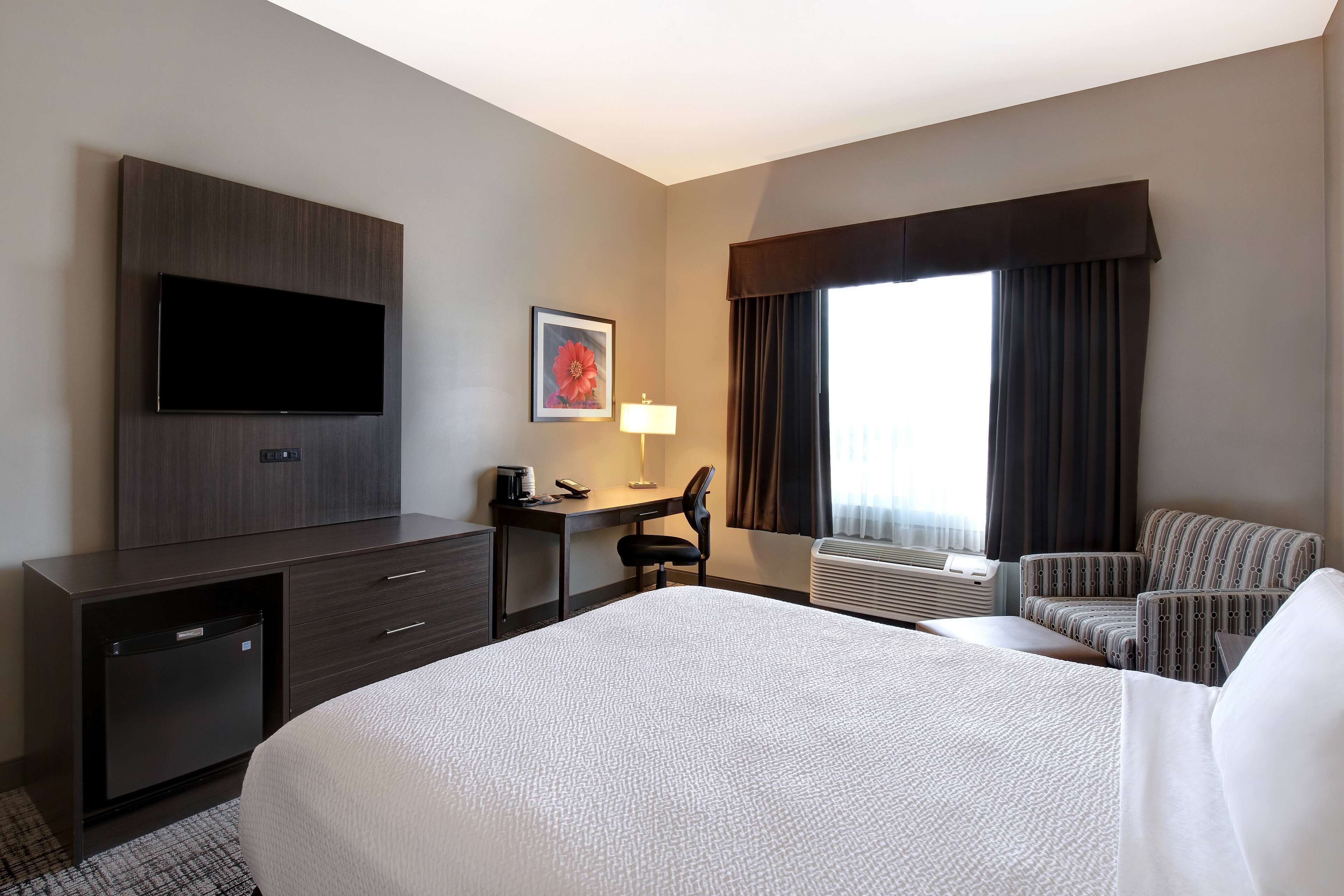 Una habitación de hotel con cama, escritorio con silla, televisor, refrigerador pequeño y ventana con cortinas.