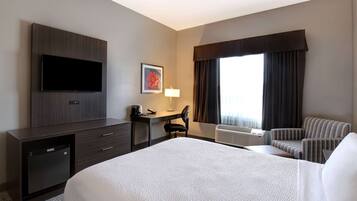 Una habitación de hotel con cama, escritorio con silla, televisor, refrigerador pequeño y ventana con cortinas.
