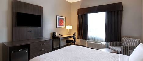 A hotel room with a bed, a desk with a chair, a television, a small refrigerator, and a window with curtains.