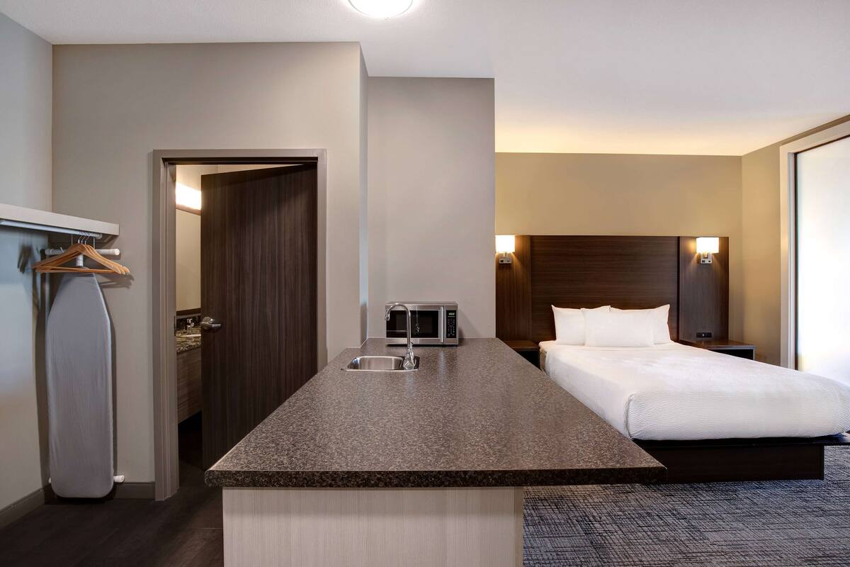 A hotel room with a kitchenette, a bed, a nightstand, and a wall-mounted lamp.