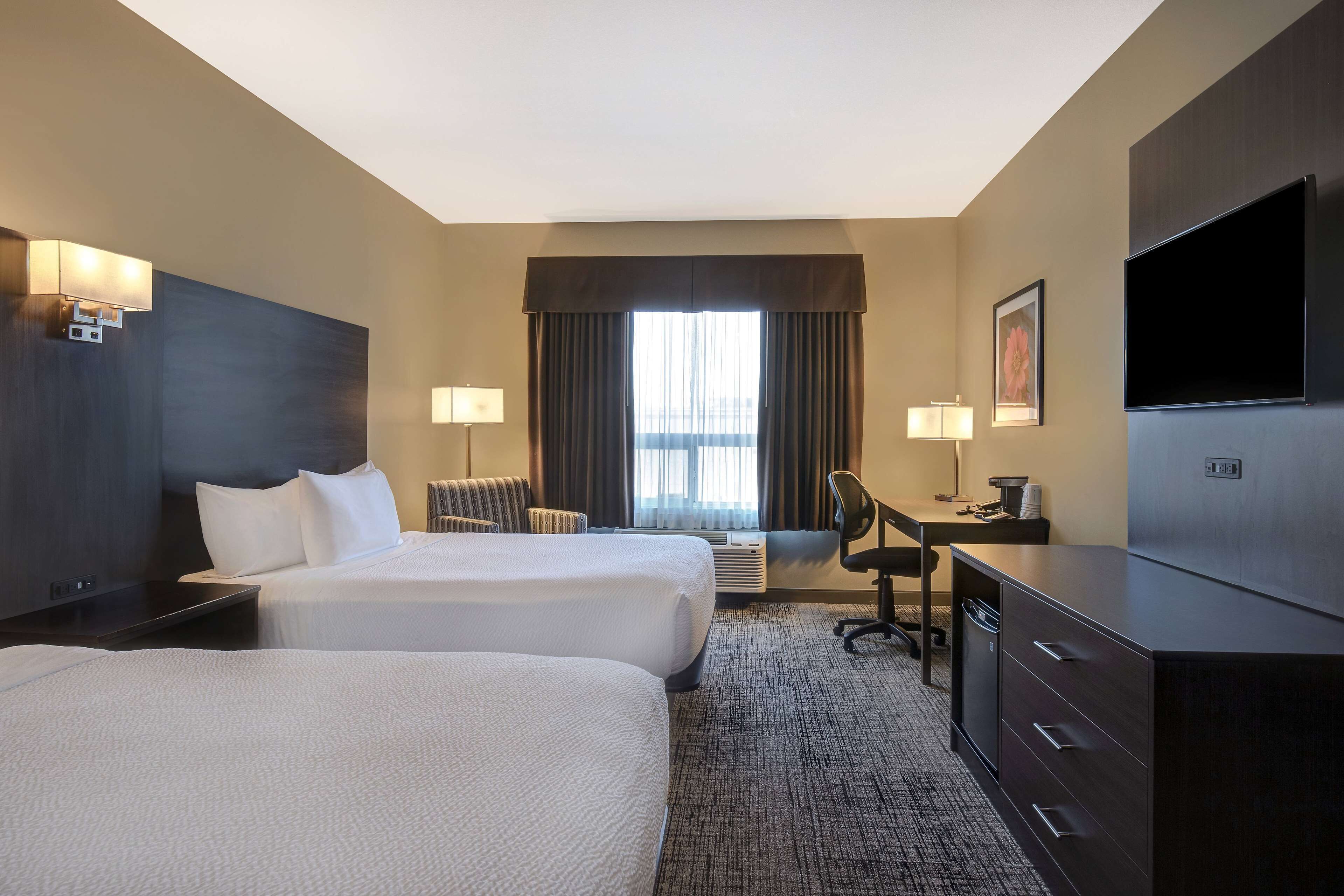 A hotel room with two beds, a desk, a chair, a TV, and a large window with curtains.