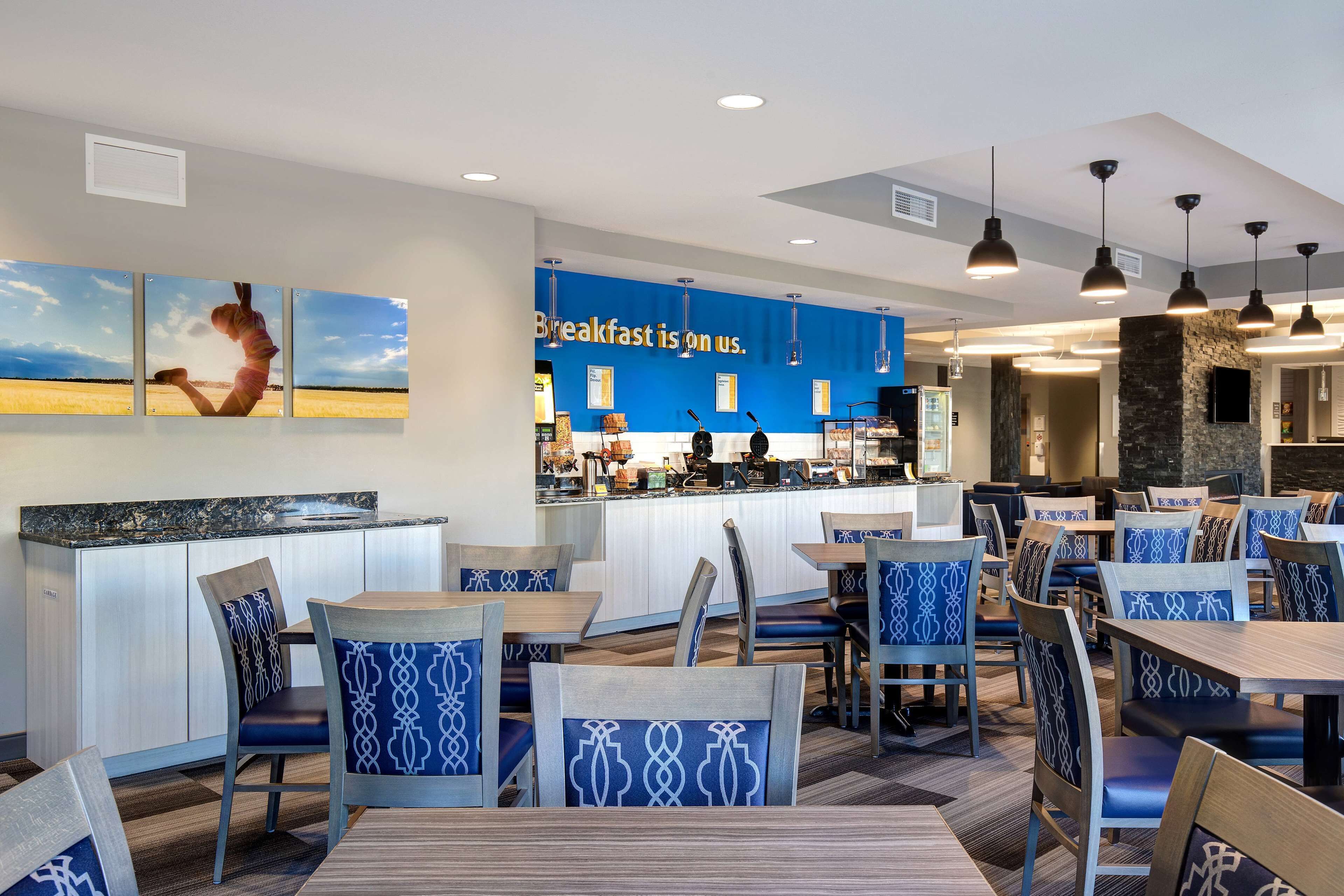 Um restaurante moderno com parede azul e um letreiro que diz "Café da manhã por nossa conta". Há várias mesas e cadeiras, e um balcão com um cardápio.