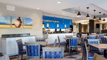 Um restaurante moderno com parede azul e um letreiro que diz "Café da manhã por nossa conta". Há várias mesas e cadeiras, e um balcão com um cardápio.