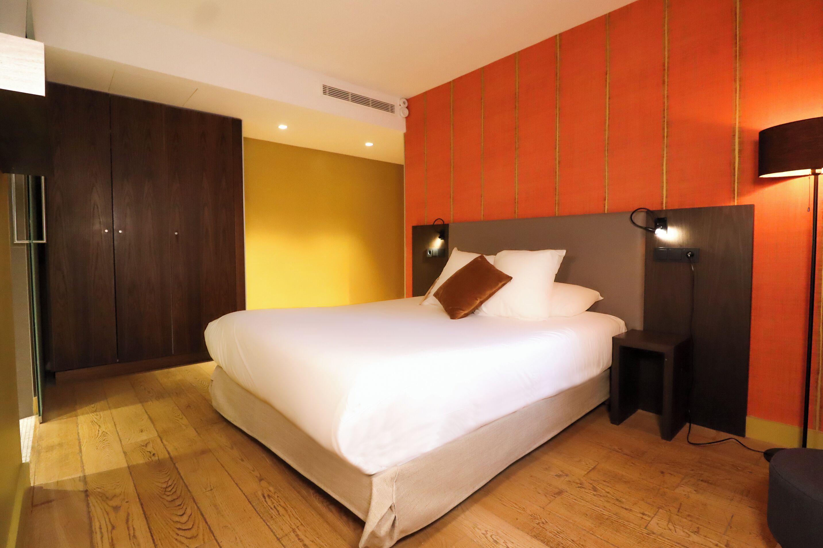 A modern hotel room with a large bed, wooden flooring, and a red accent wall.