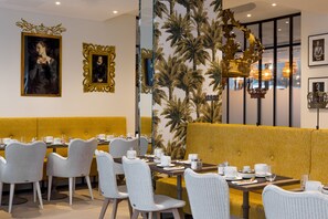 A restaurant with white wicker chairs, yellow cushioned booths, and framed artwork on the walls. - Hotel l'Arbre Voyageur (Lille)