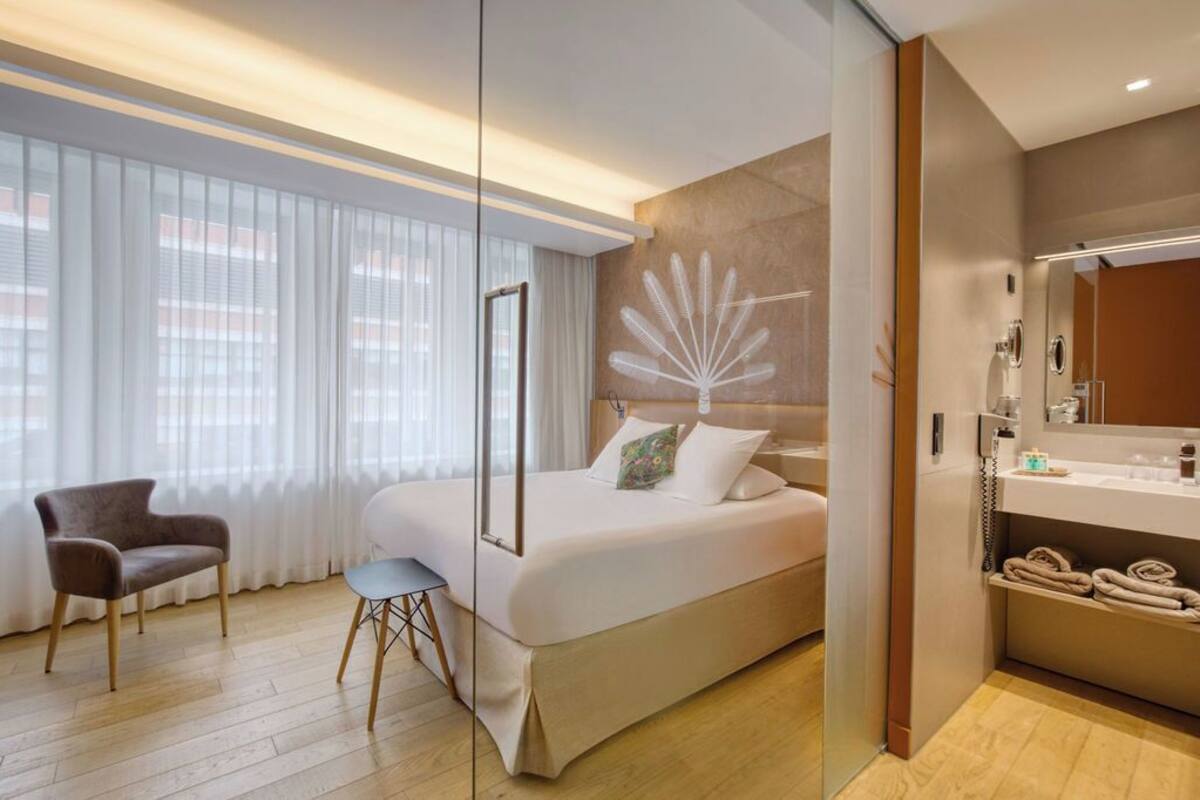 Ein modernes Hotelzimmer mit einem großen Bett, einem Sessel, einem Schreibtisch und einem Badezimmer mit Spiegel und Handtuchhalter.