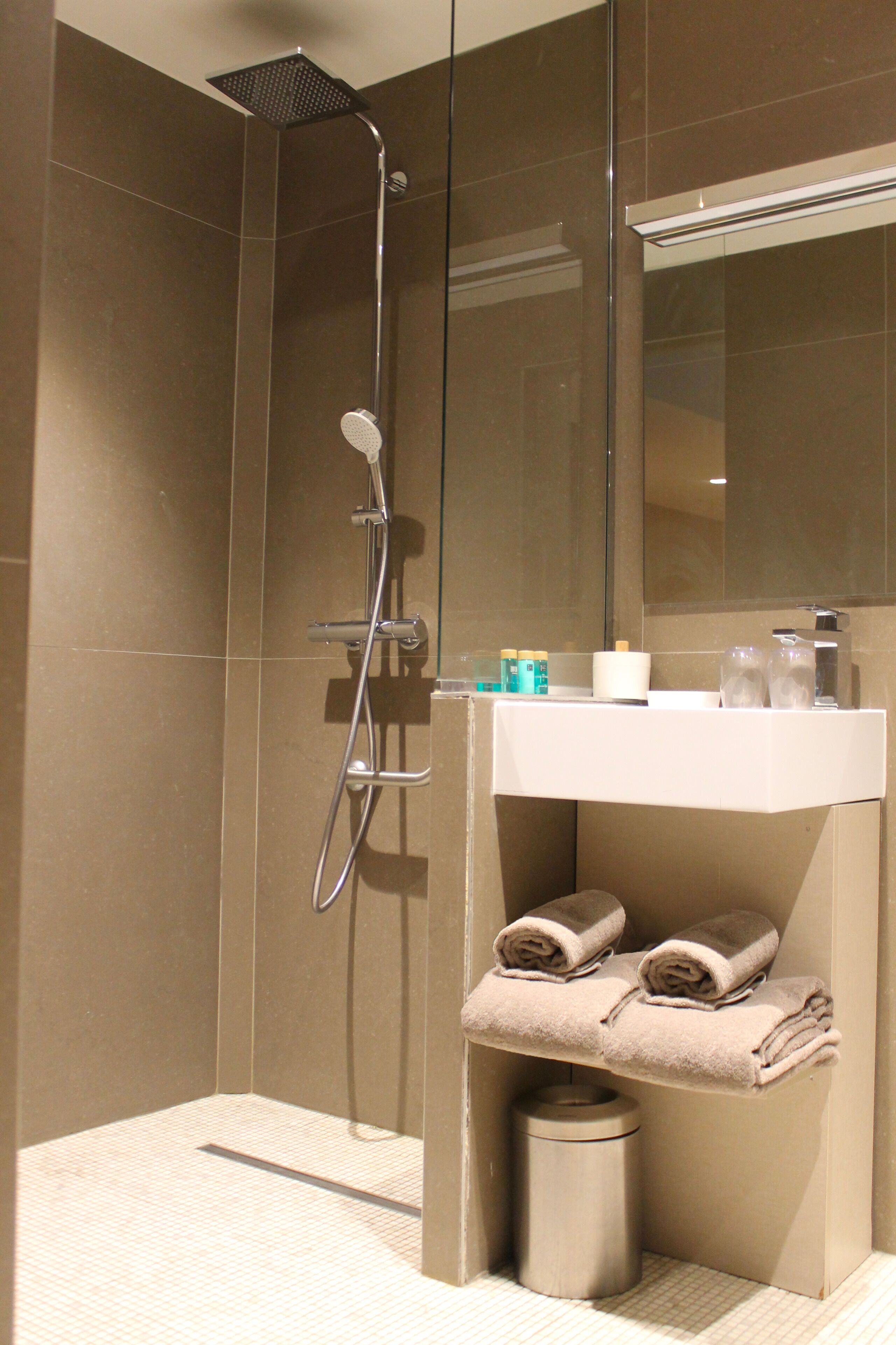 Une salle de bain moderne avec une douche à parois de verre, un lavabo blanc avec un miroir et des serviettes soigneusement pliées sur une étagère.