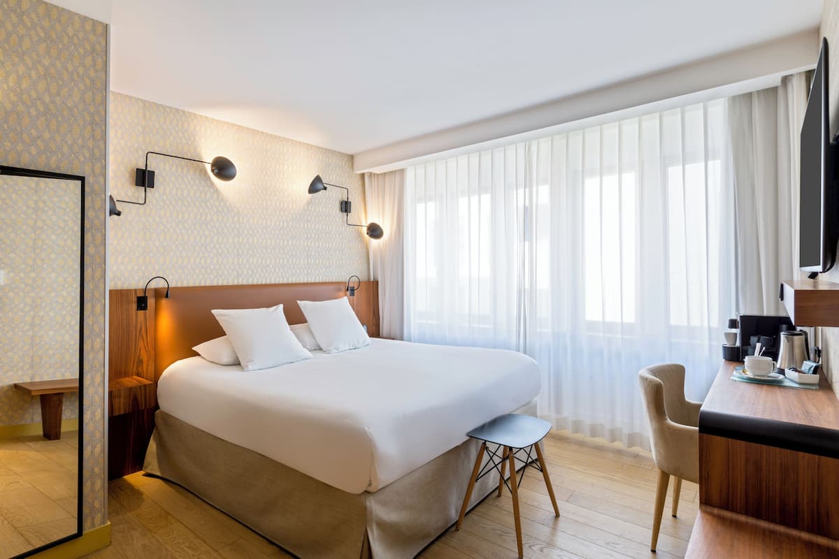 Ein Hotelzimmer mit einem großen Bett, Nachttischen, einem Schreibtisch mit Stuhl, einem Fernseher und einem fenster mit Vorhängen.