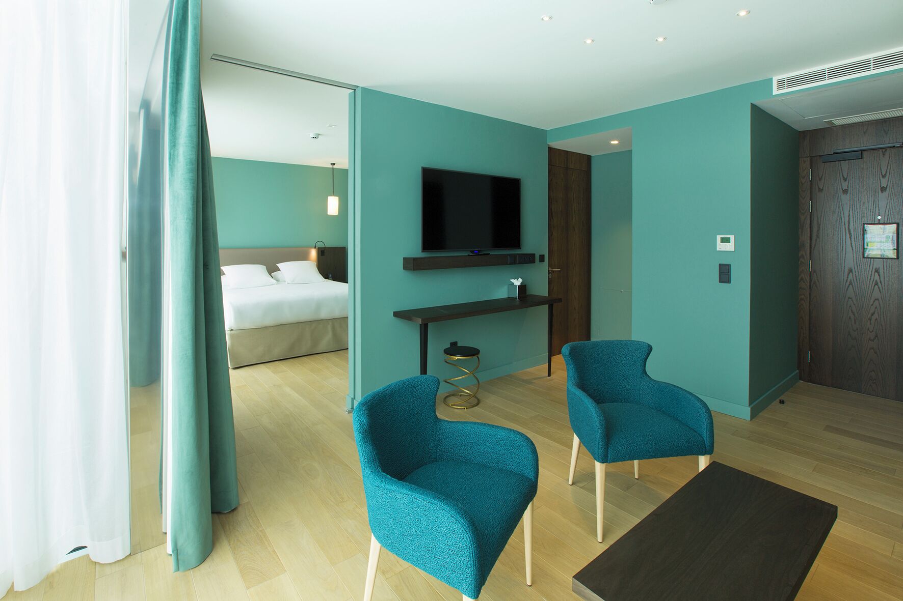Een moderne hotelkamer met een bed, een aan de muur bevestigde televisie, twee blauwgroene stoelen en een houten vloer.