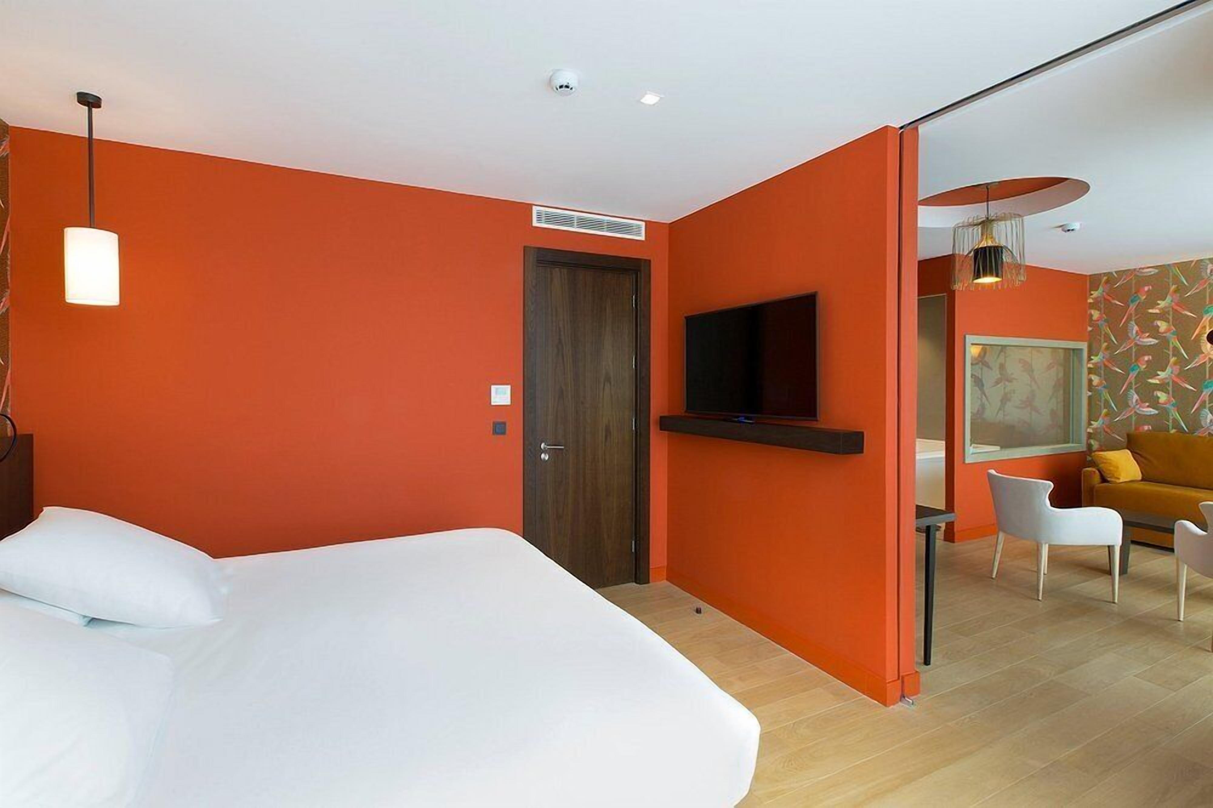 A modern hotel room with an orange wall, a large bed, a TV mounted on the wall, a sofa, and a chair.