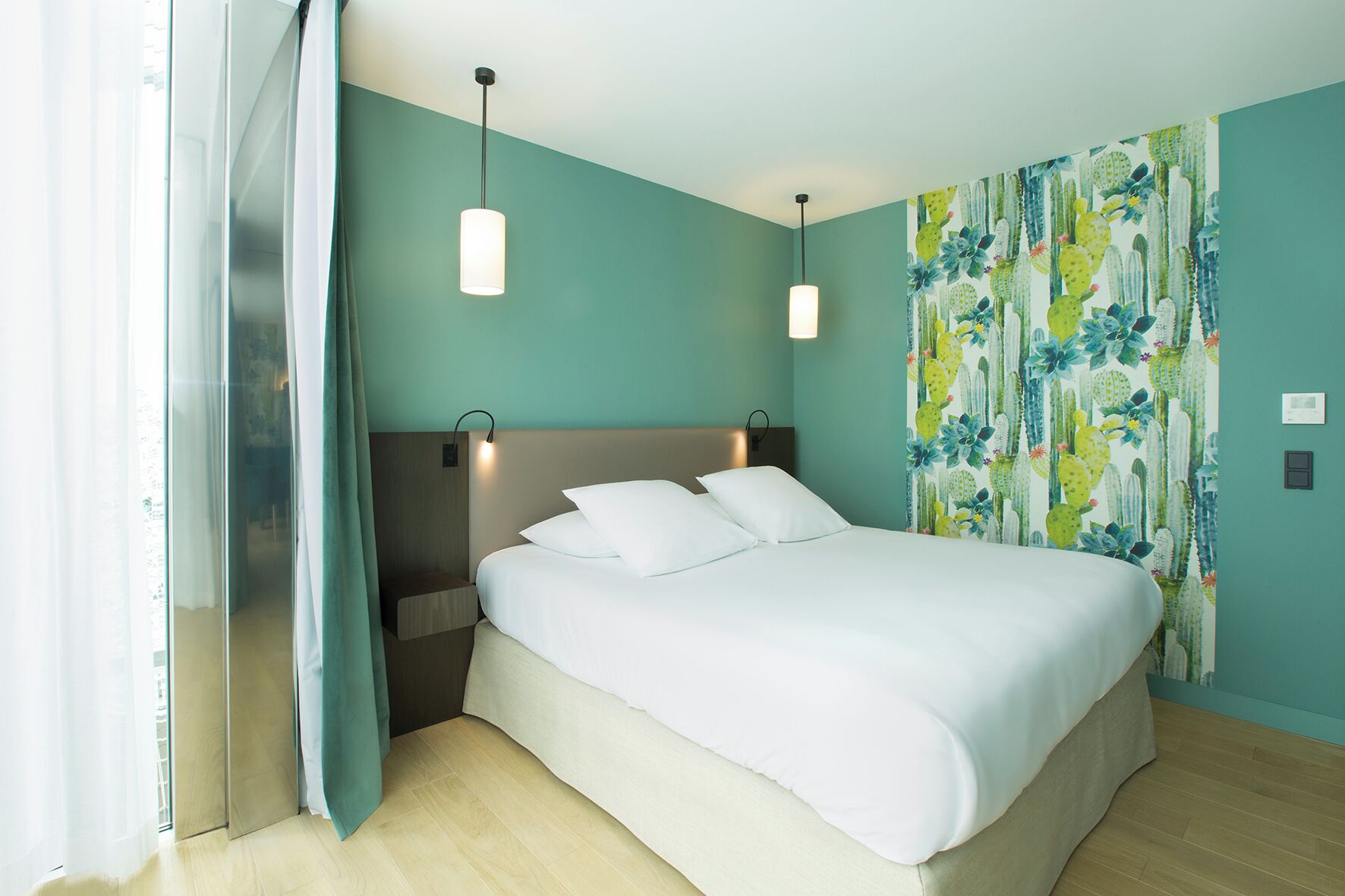 A modern hotel room with a large bed, two bedside lamps, a nightstand, and a cactus-themed wall.