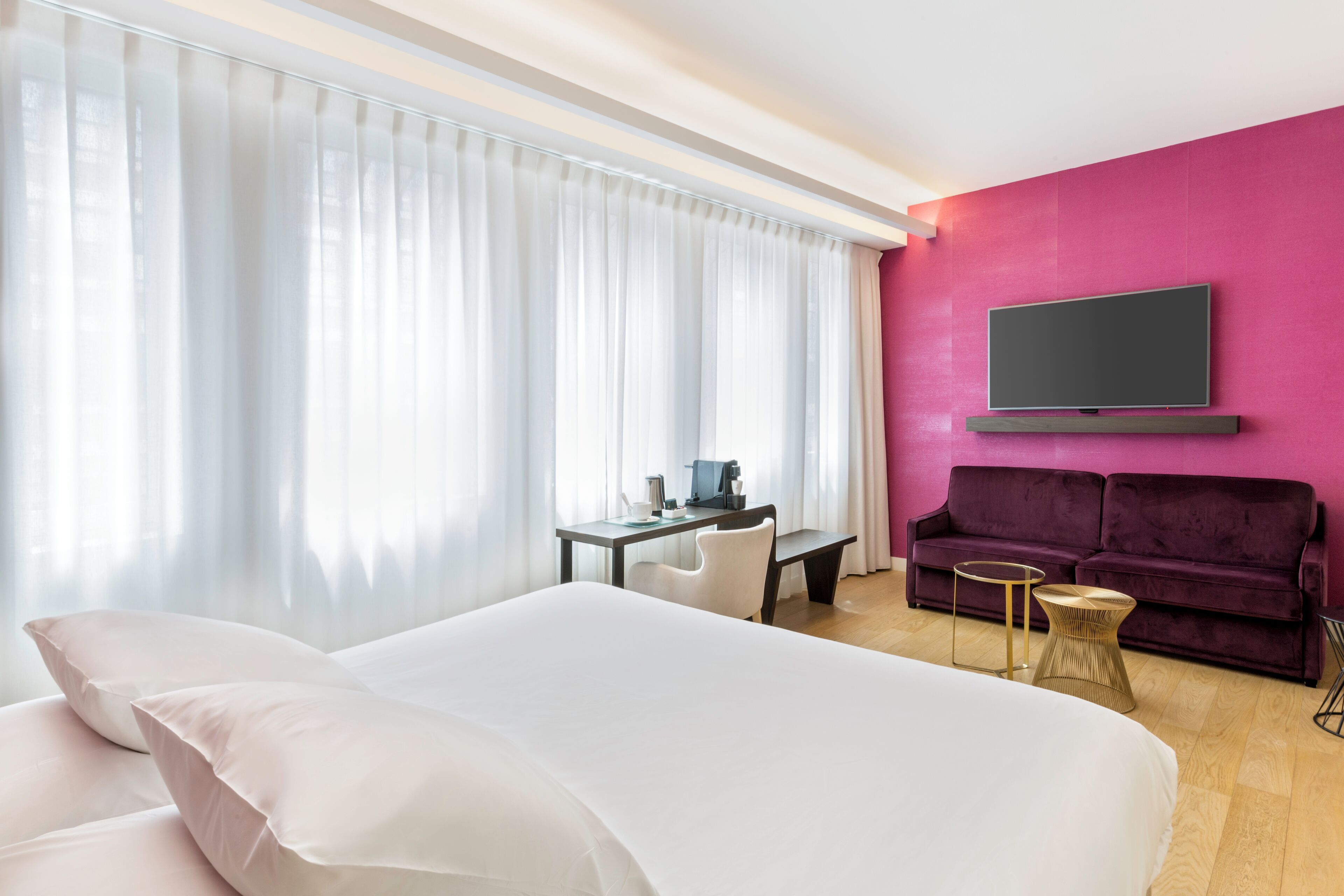 A hotel room with a large bed, a desk with a chair, a purple sofa, and a TV mounted on the wall.