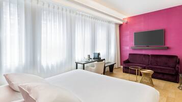 A hotel room with a large bed, a desk with a chair, a purple sofa, and a TV mounted on the wall.