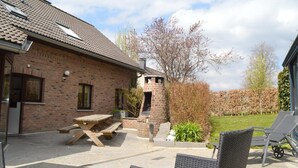 A brick house with a tiled roof, a patio with a wooden table and chairs, and a garden with a lawn and trees. - Holiday Home in Waimes with Pool & Sauna (Waimes)