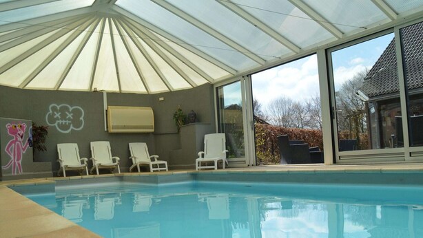 An indoor swimming pool with a glass ceiling, white lounge chairs, and a large window. - Holiday Home in Waimes with Pool & Sauna (Waimes)