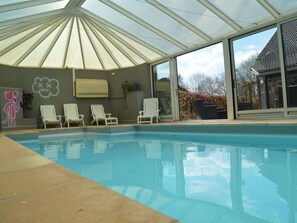 An indoor swimming pool with a glass ceiling, white lounge chairs, and a large window.