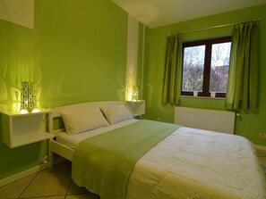Un dormitorio con cama, mesitas de noche, una ventana con cortinas y un radiador.