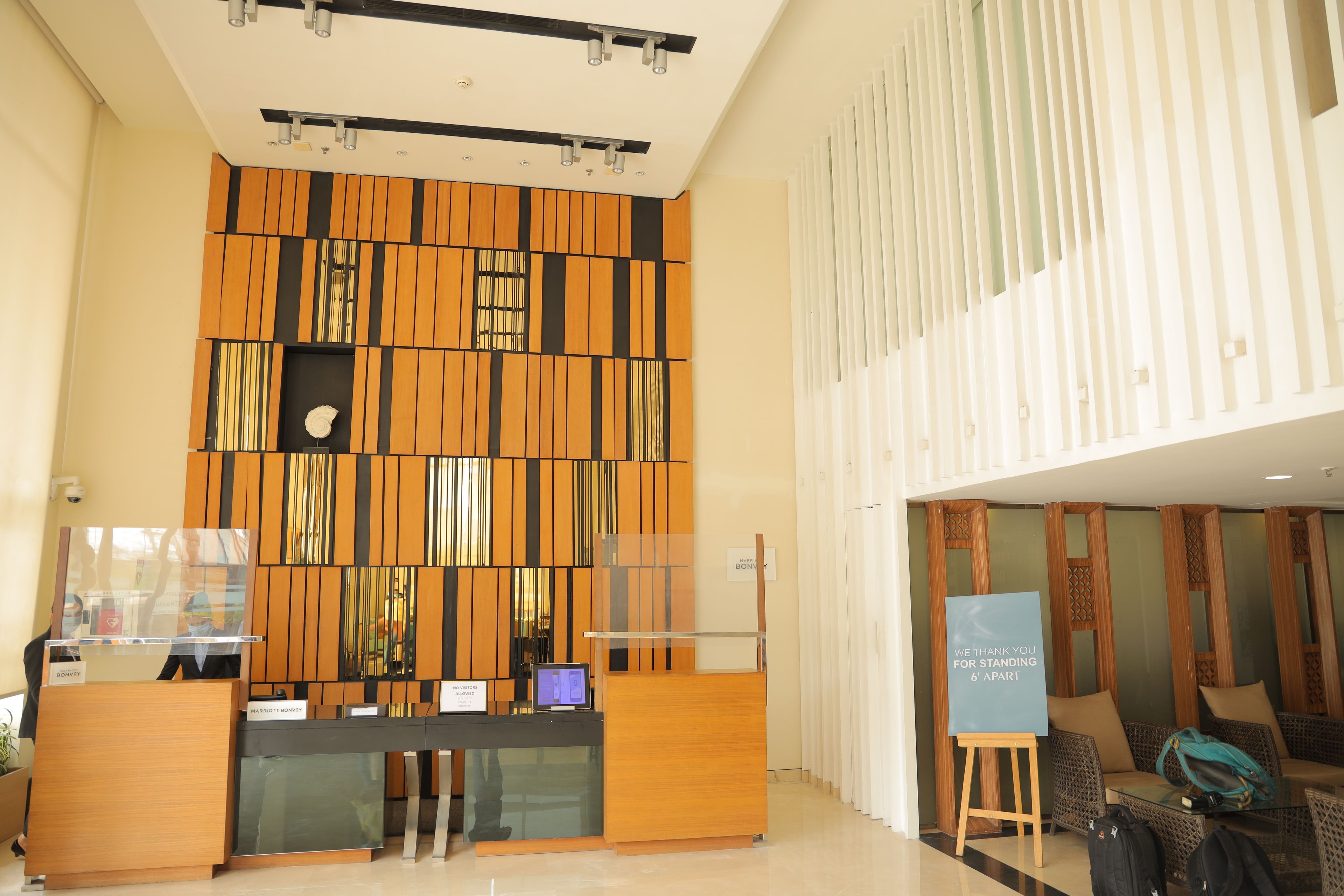 Egy modern dizájnnal megtervezett szállodai recepciós tér, mely tartalmaz egy recepciós pultot, egy digitális kijelzőt és egy ülősarokot, ahol egy tábla fejezi ki a vendégek iránti hálát a türelmükért.