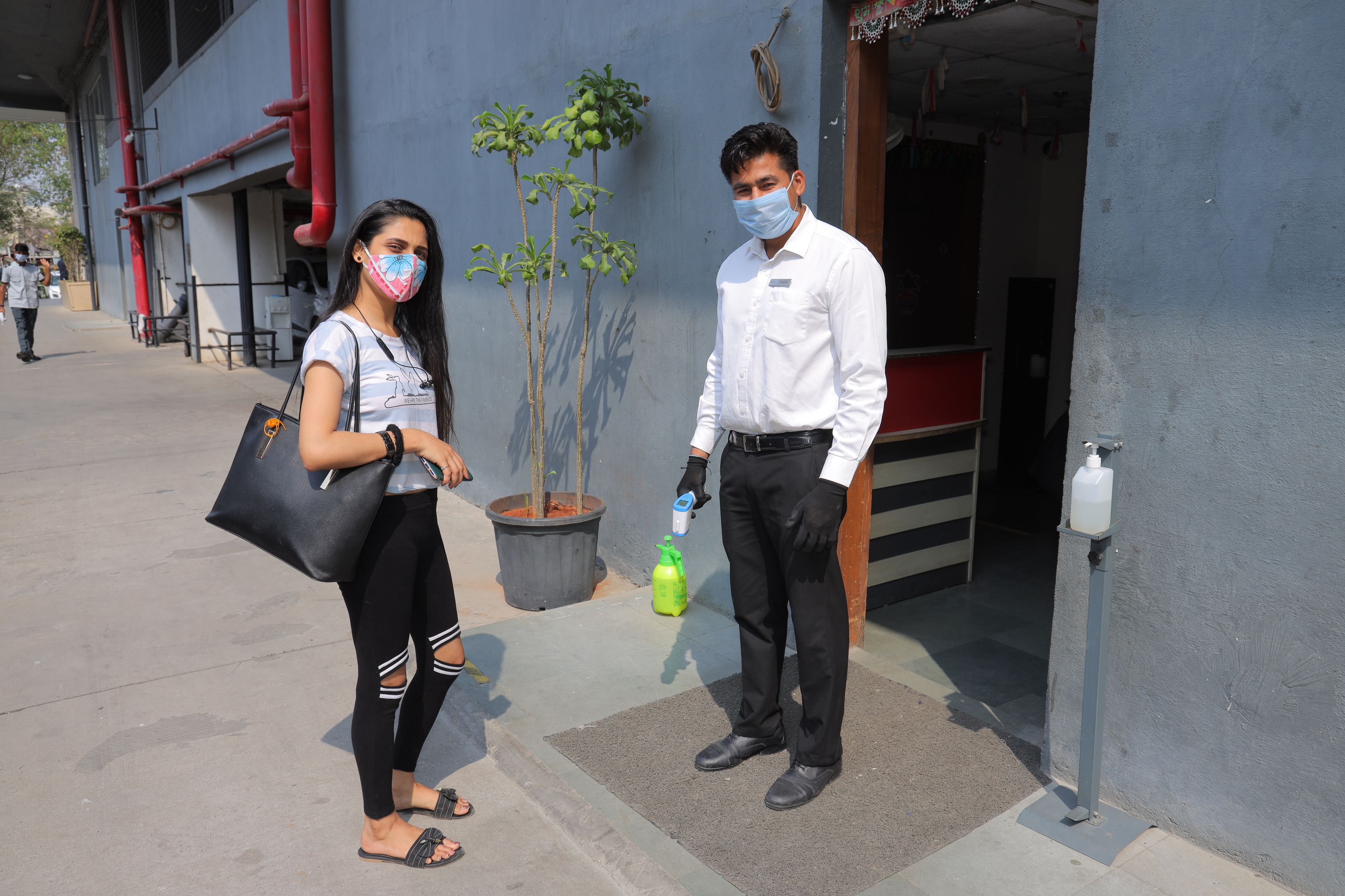 Two individuals standing outside a building with a grey exterior, a potted plant, and a utility box.