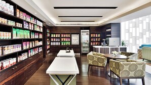 Un moderno vestíbulo de hotel con un mostrador de recepción alargado, zona de estar y varios productos envasados en estanterías.