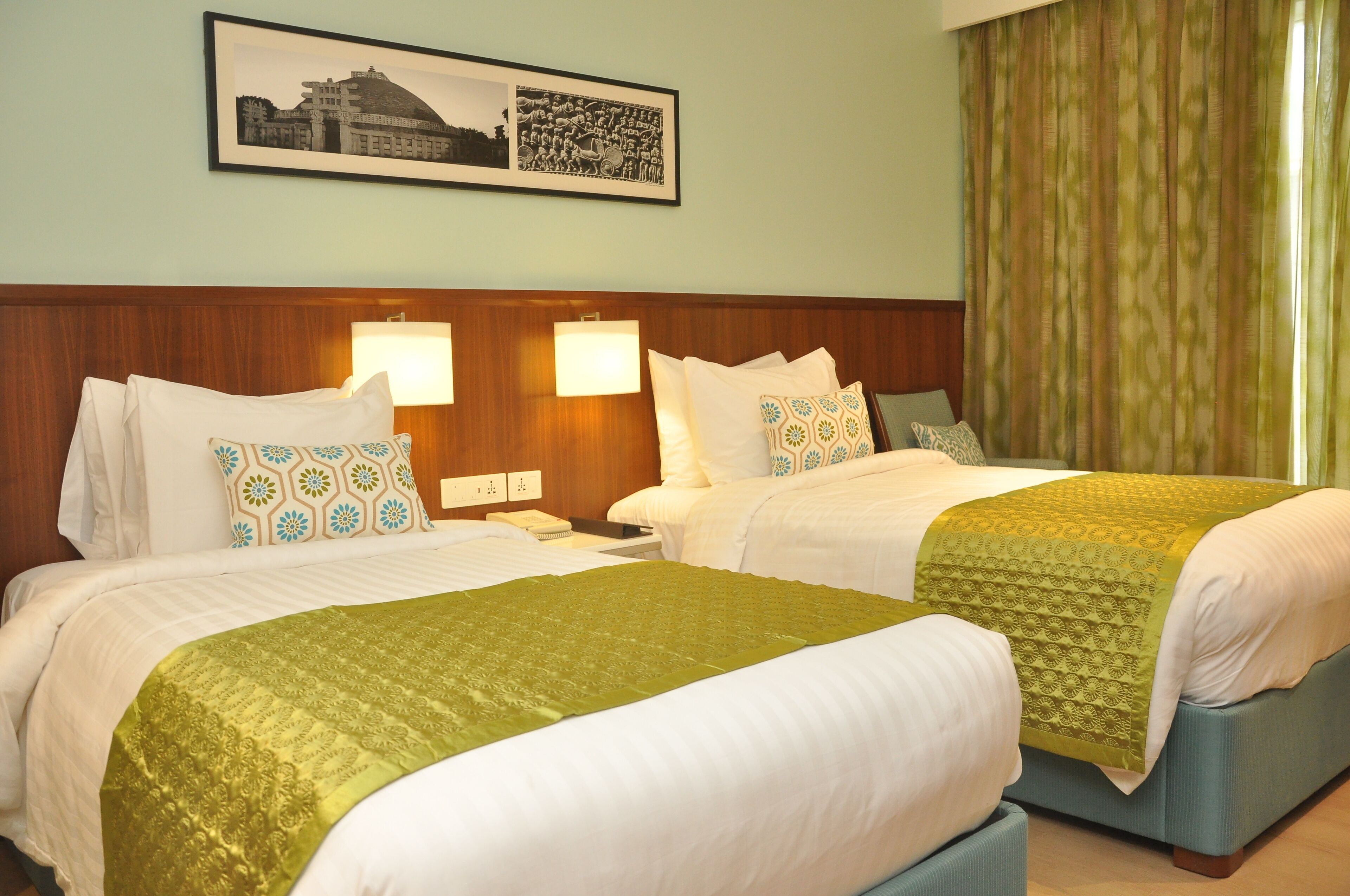 A hotel room with two beds, each with patterned pillows and a green bedspread. There are wall-mounted lamps and a framed picture above the beds.