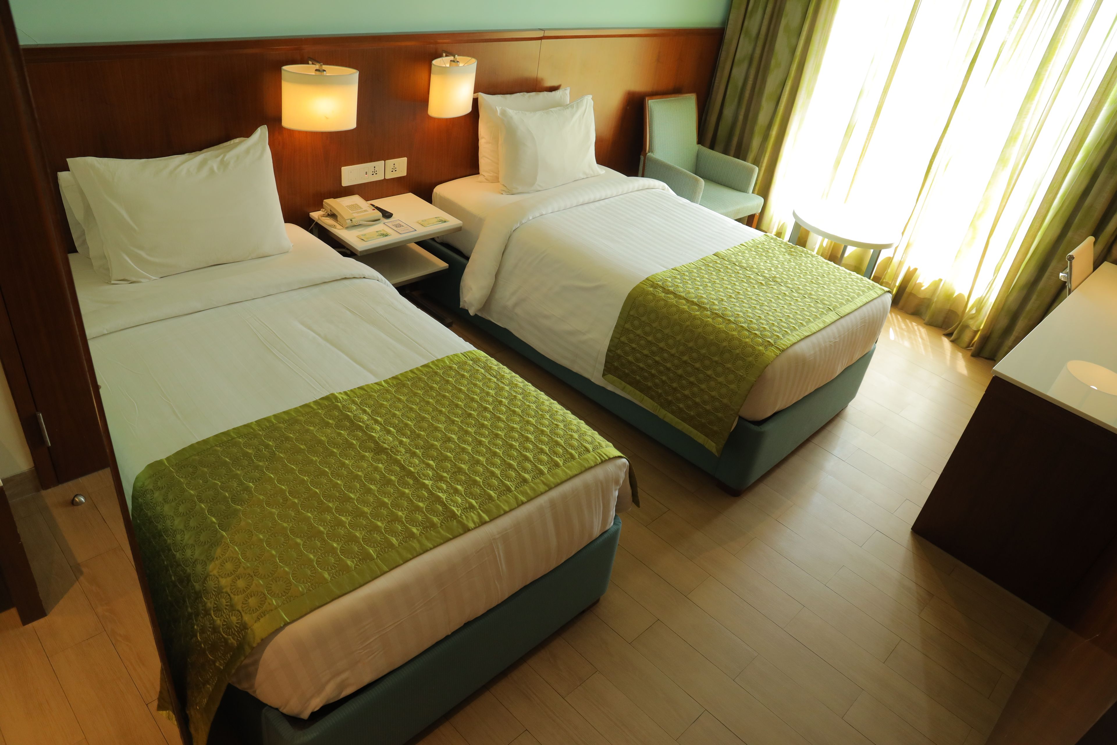 A hotel room with two beds, a bedside table with a lamp, a telephone, and a window with curtains.
