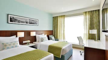 A hotel room with two beds, a desk, and a chair. There is a window with curtains and a framed picture on the wall.