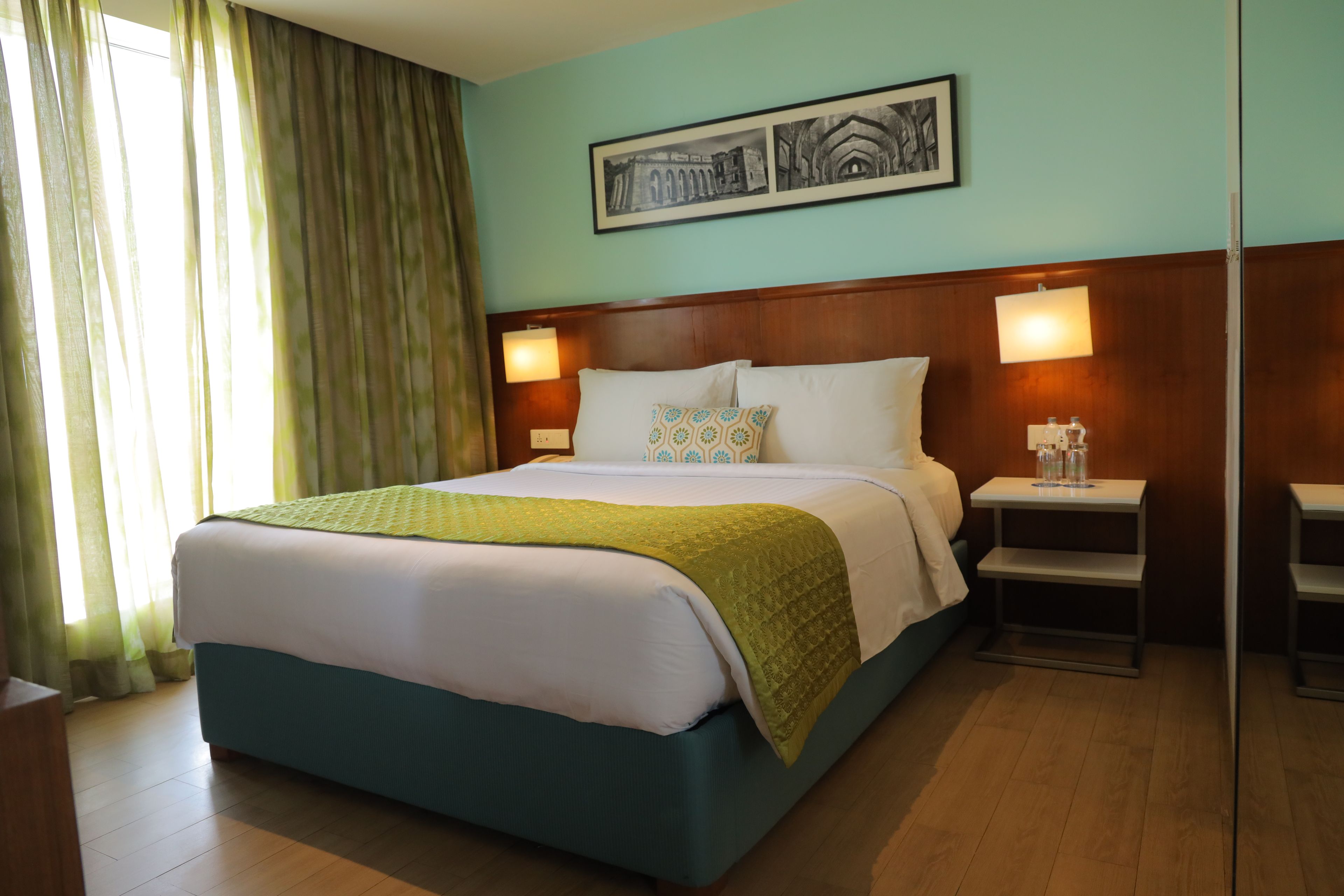 Une chambre d’hôtel avec un grand lit, deux tables de chevet, une commode, une fenêtre avec des rideaux et un tableau encadré au mur.