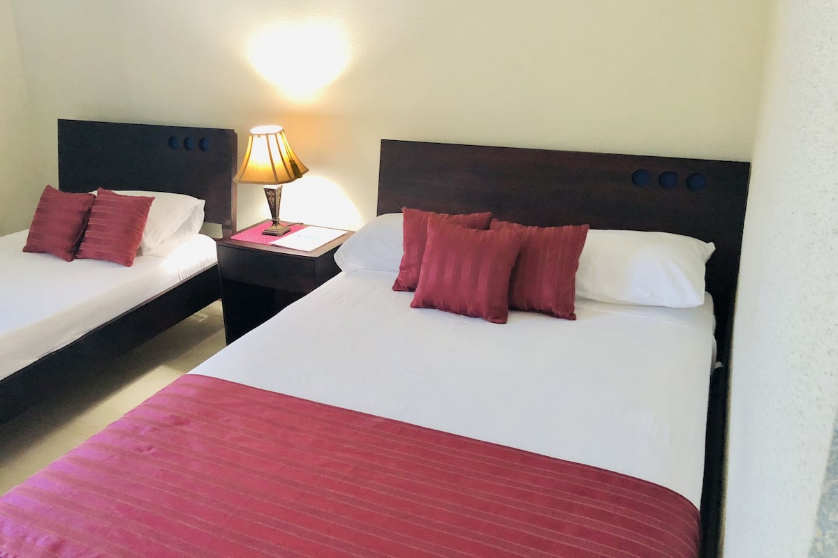Une chambre d’hôtel avec deux lits simples, chacun agrémenté d’un oreiller rouge et de draps blancs. Une table de chevet avec une lampe se trouve entre les deux lits.
