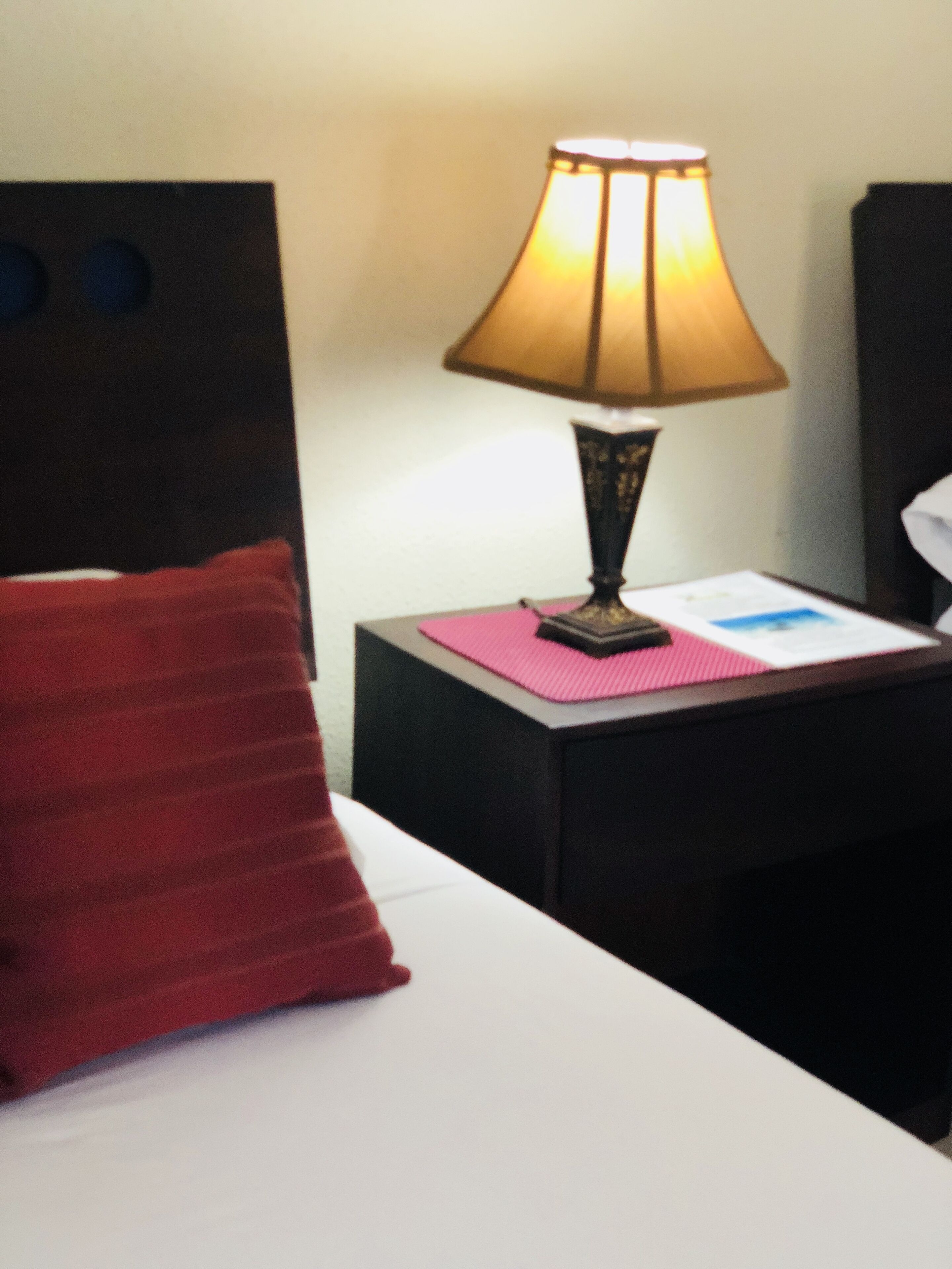 A hotel room with a bed, a nightstand with a lamp, a red pillow, and a pink placemat.