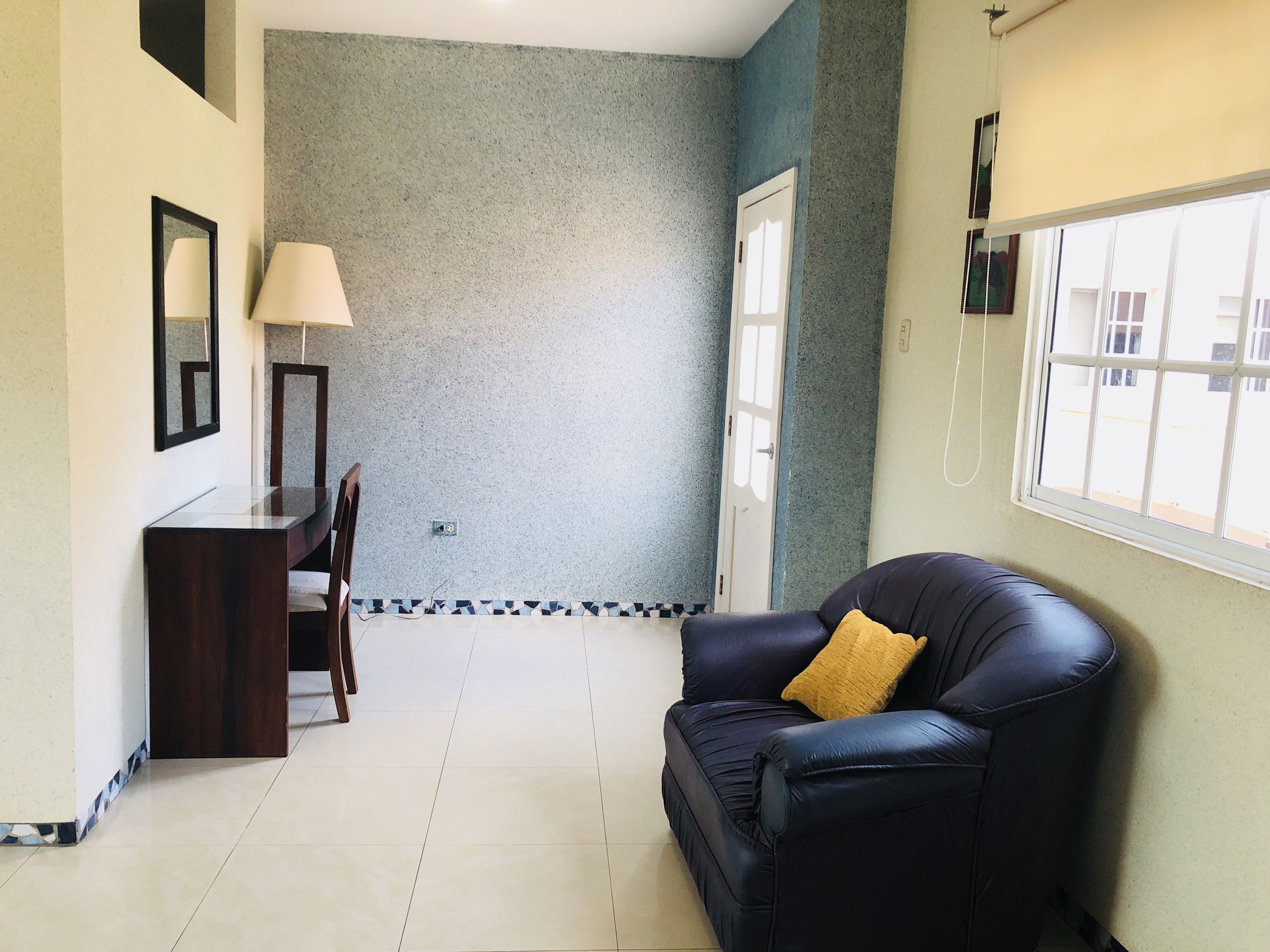 Una habitación con un sofá de cuero oscuro, un escritorio pequeño con lámpara, suelo de baldosas y ventana con vistas.