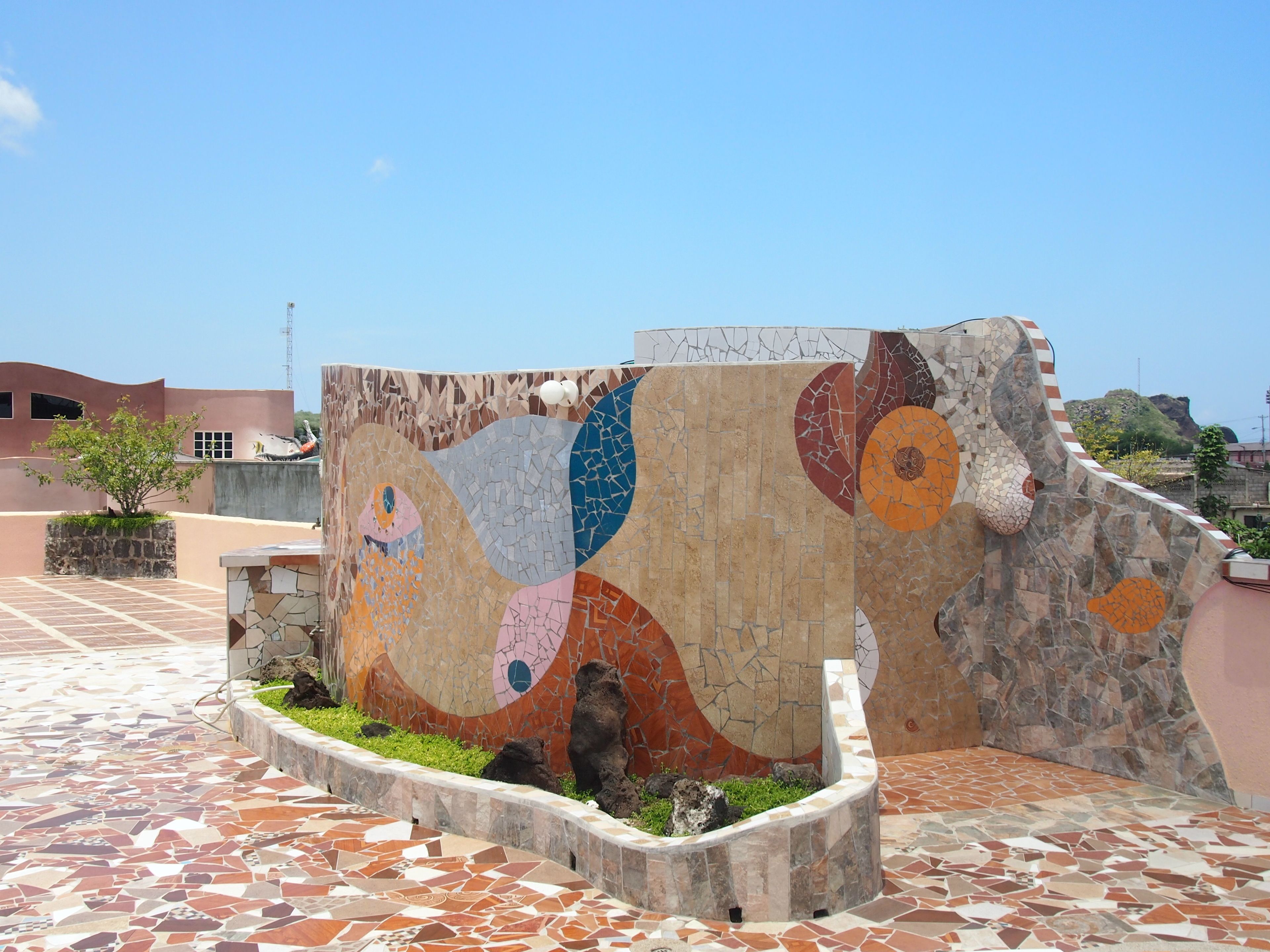 Una pared de mosaicos con patrones abstractos, un patio empedrado y una jardinera de piedra con plantas.