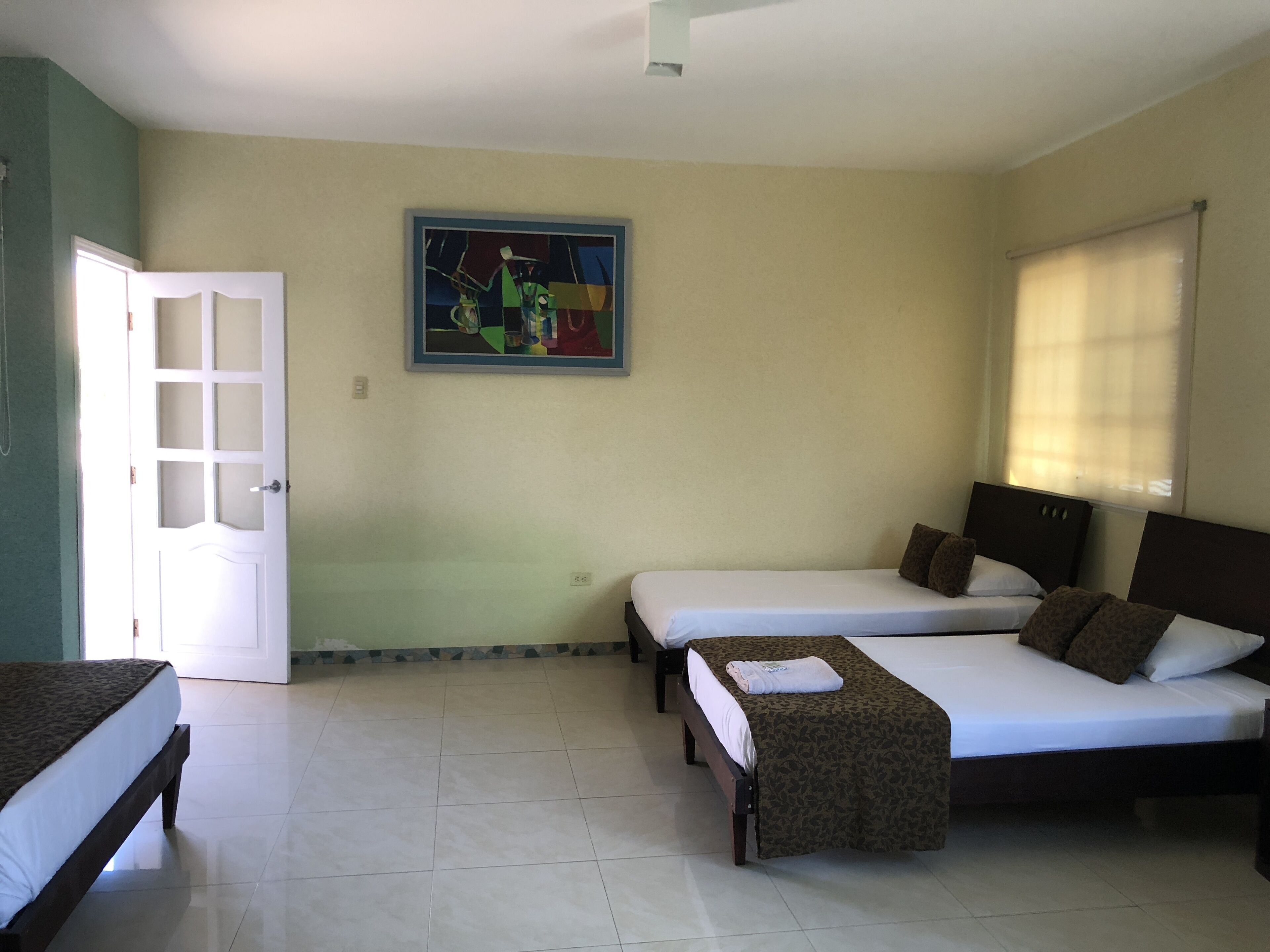 A hotel room with two single beds, a large window with blinds, a door with a frosted glass panel, and a painting on the wall.