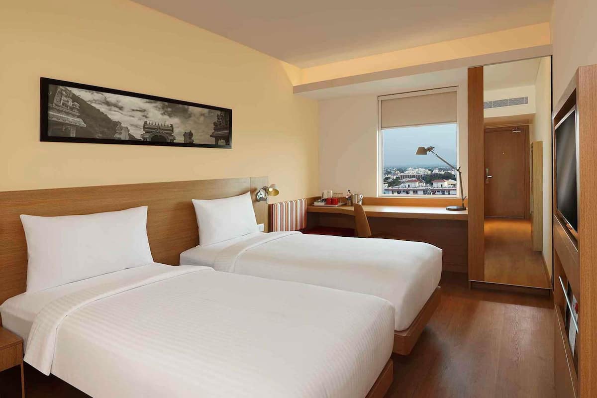 A hotel room with two beds, a desk, a chair, a TV, and a window with a view.