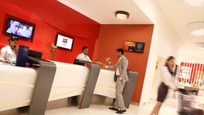 Zona de recepción de un hotel con pared roja, mostrador de recepción, pantalla de televisión y pantalla digital.