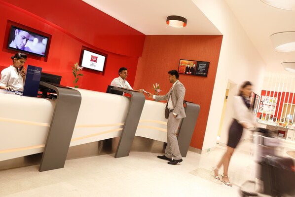 Zona de recepción de un hotel con pared roja, mostrador de recepción, pantalla de televisión y pantalla digital.