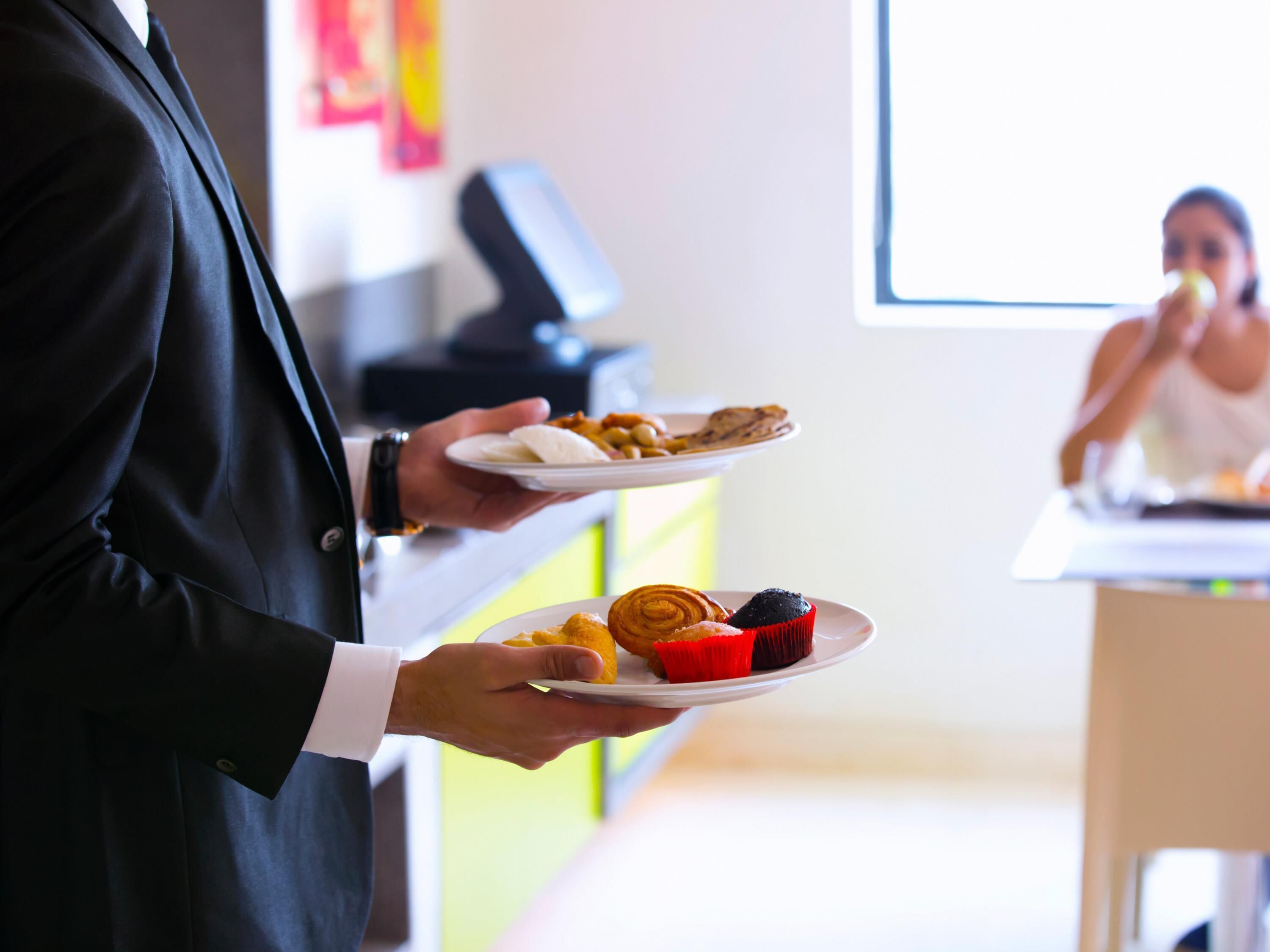 A person holding plates with food items, a counter, and a blurred figure in the background.