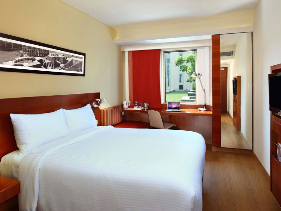 Habitación de hotel con cama, escritorio, silla, televisor, ventana que ofrece vistas a edificios y follaje, y un cuadro enmarcado en la pared.