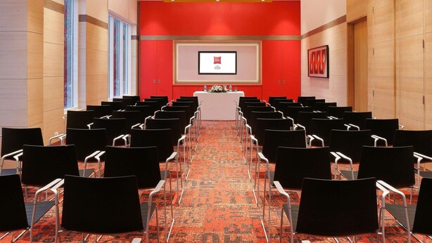 Un salón de conferencias con filas de sillas, un atril y una pantalla mostrando un logotipo.