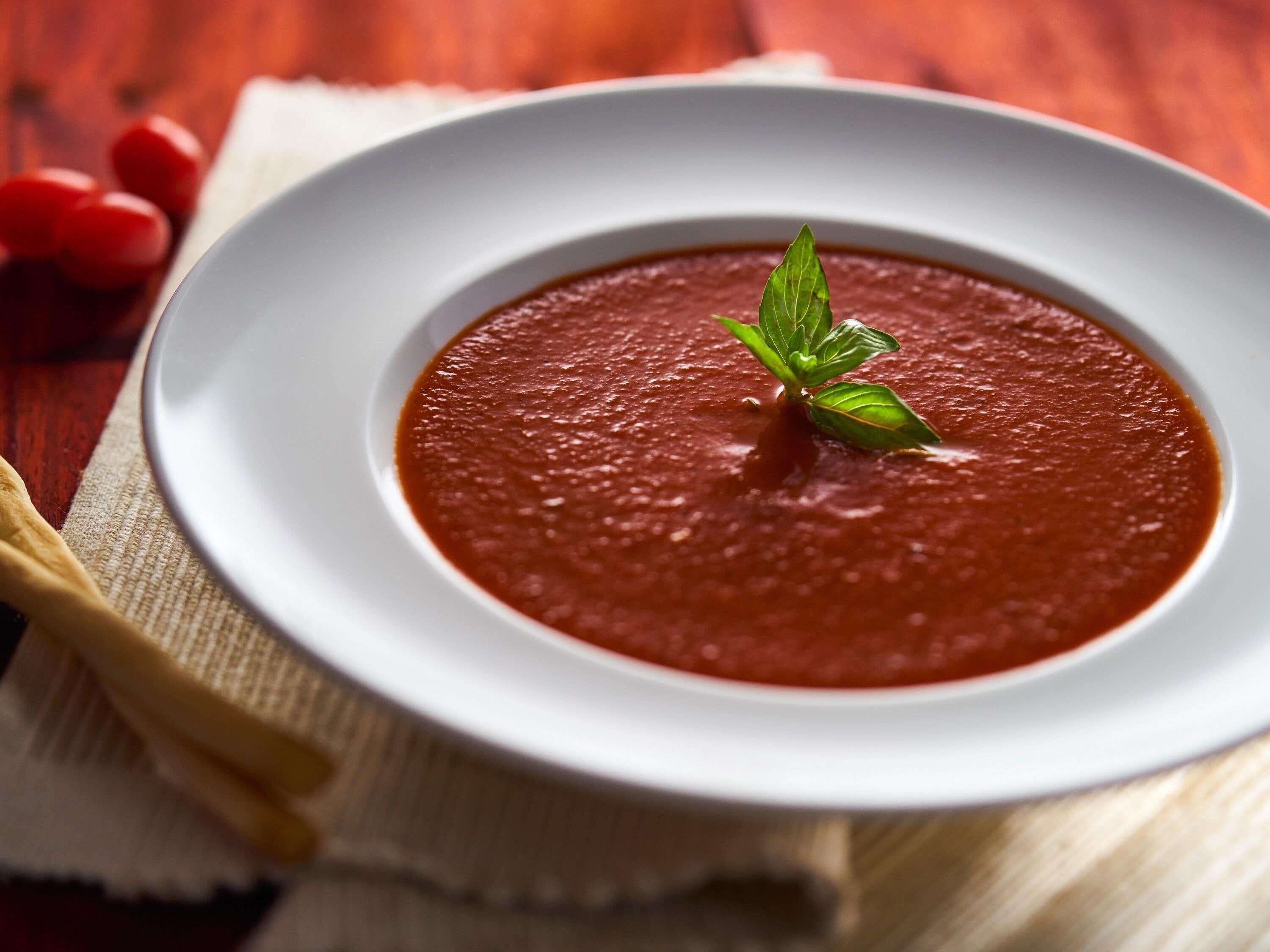 A bowl of tomato soup garnished with a basil leaf on a white plate.