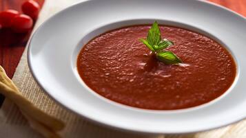 Un plato blanco con un tazón de sopa de tomate adornado con una hoja de albahaca.